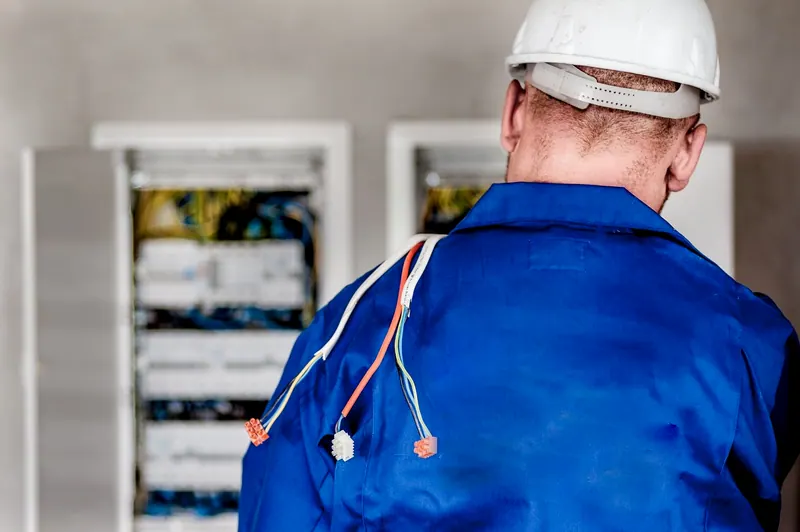 Eletricista de uniforme azul diante de quadros de distribuição abertos